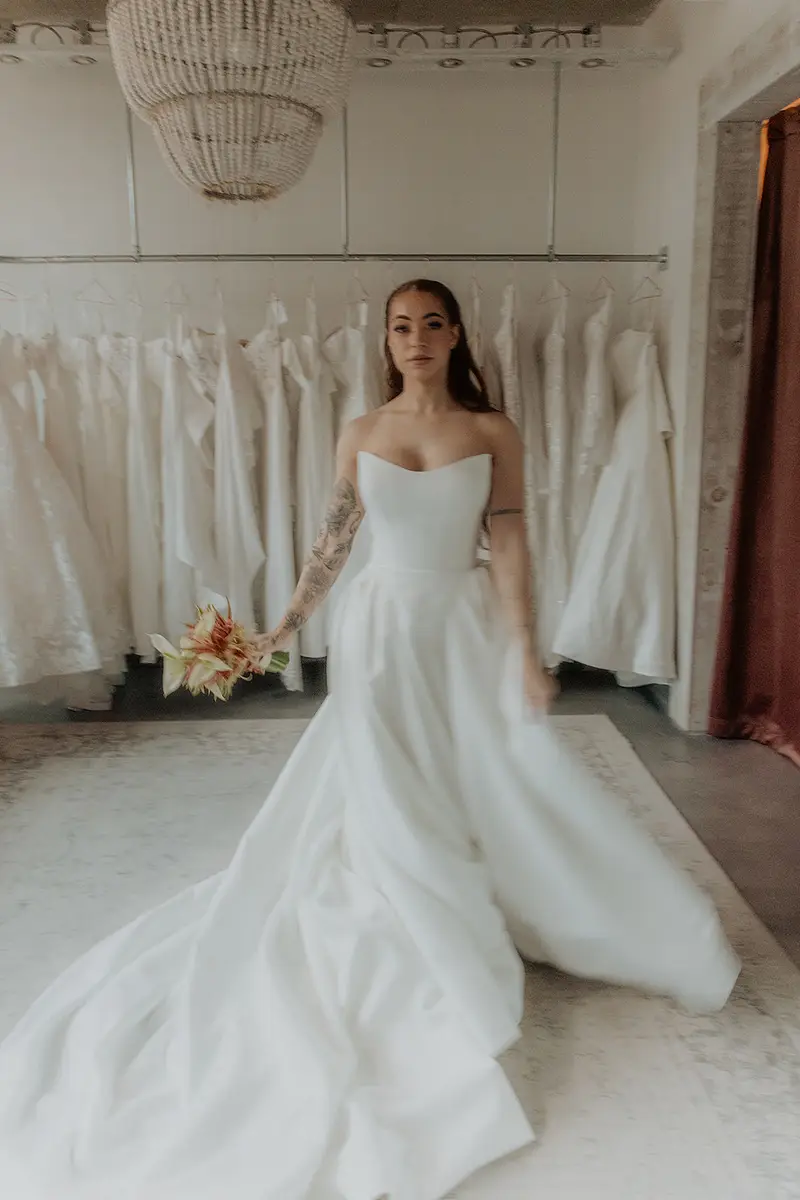 Bride in a strapless Private Collection ballgown with a plain skirt and a long train, in front of the gown rail and beaded chandelier at Cande Bridal Boutique