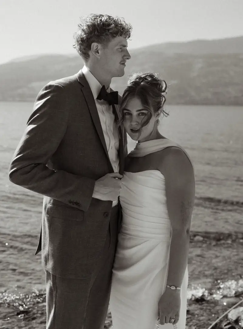Black and white photograph of a bride in a draped satin gown standing with her husband on the shore of Okanagan Lake
