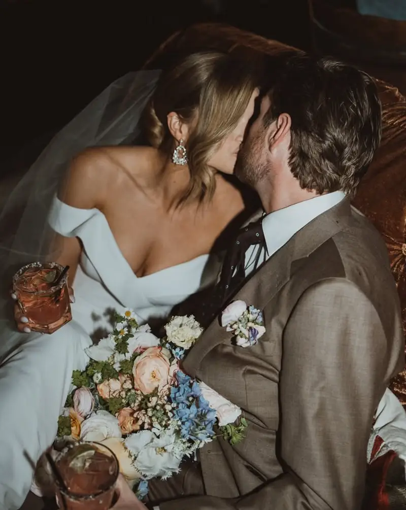 Bride in an off-the-shoulder gown leaning into her husband on a velvet sofa at the reception, cocktails and a garden bouquet in front of them