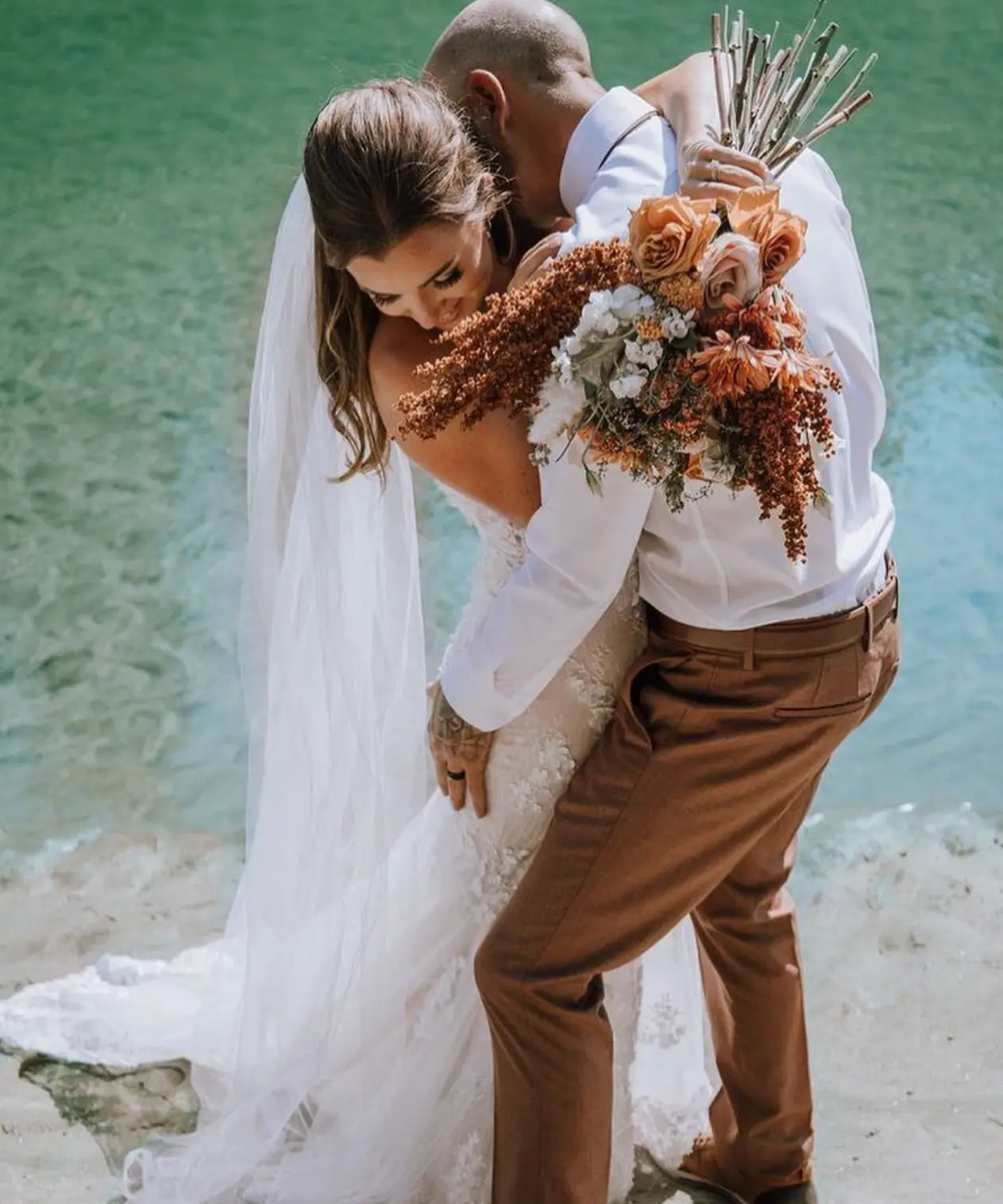 A bride and her partner kissing beside a lake on their wedding day, in a gown sold by Cande Bridal Boutique in Kelowna, British Columbia