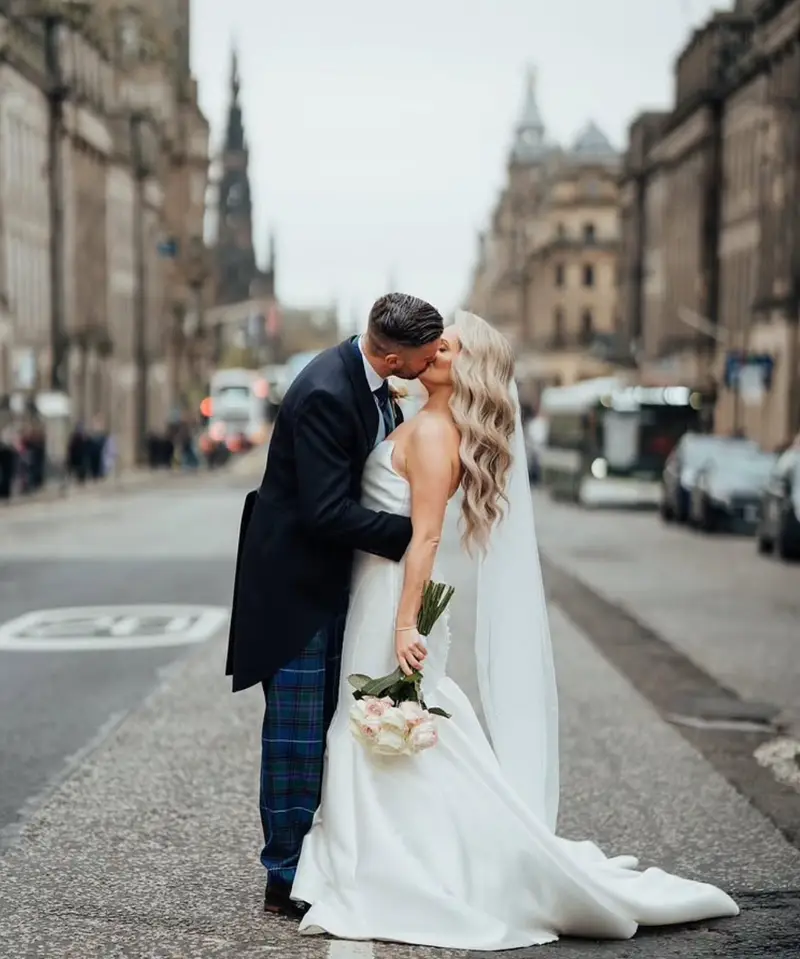 Bride and groom kissing in the middle of an Edinburgh street, the bride in a plain satin gown and long veil holding pale roses, the groom in tartan trousers