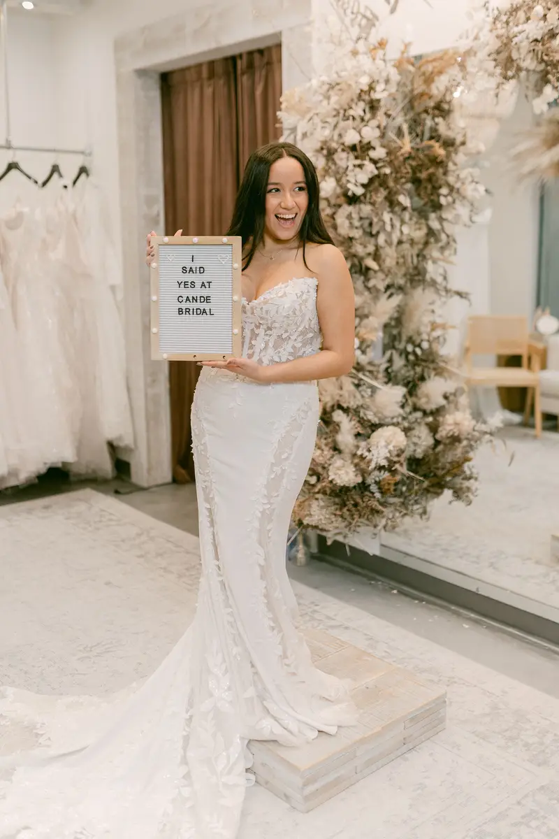 Bride in a lace fit-and-flare gown on the fitting platform at Cande Bridal Boutique, holding a letter board that reads I said yes at Cande Bridal